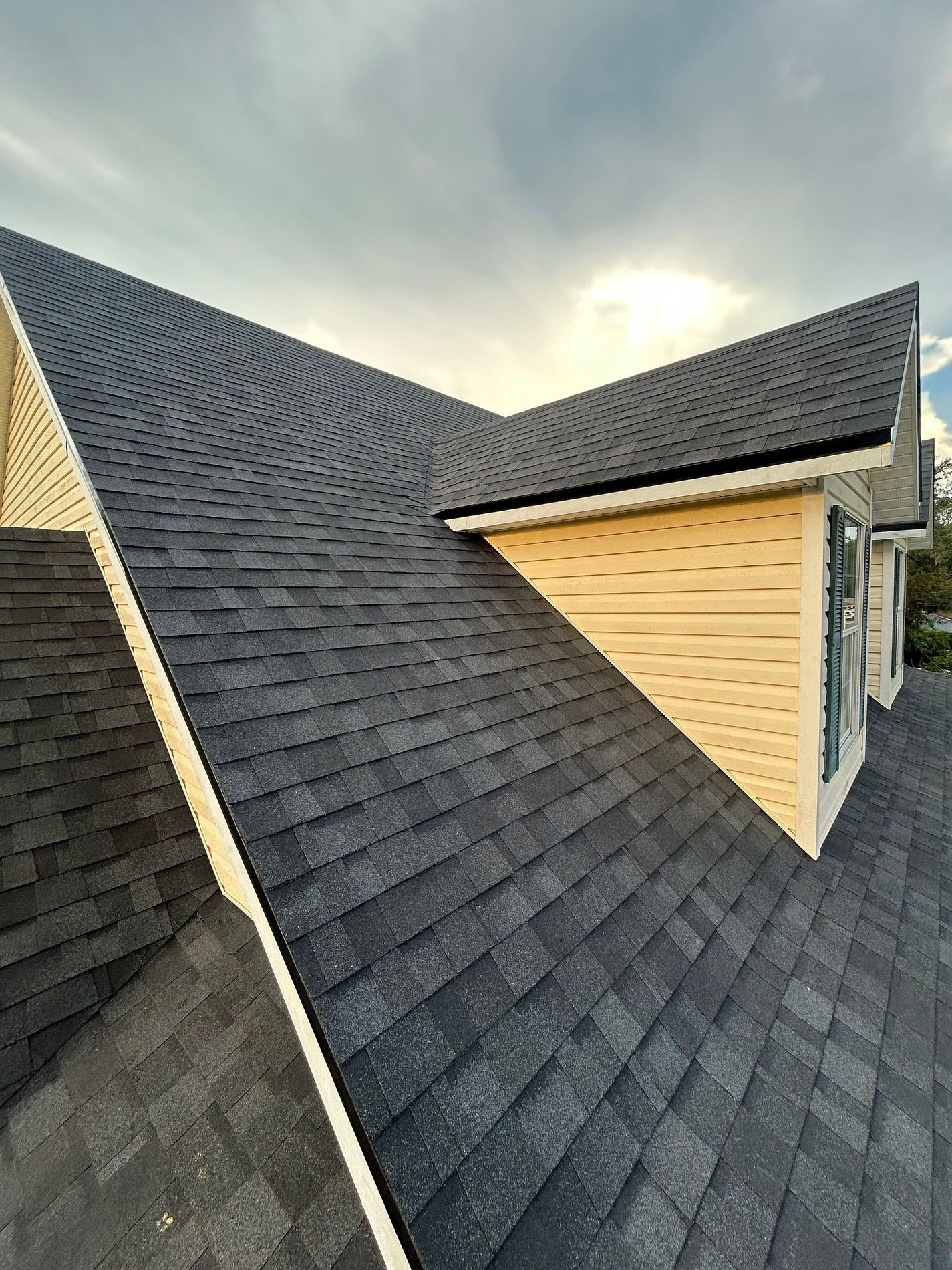 Dark shingled roof of a house with light tan siding and a cloudy sky background.