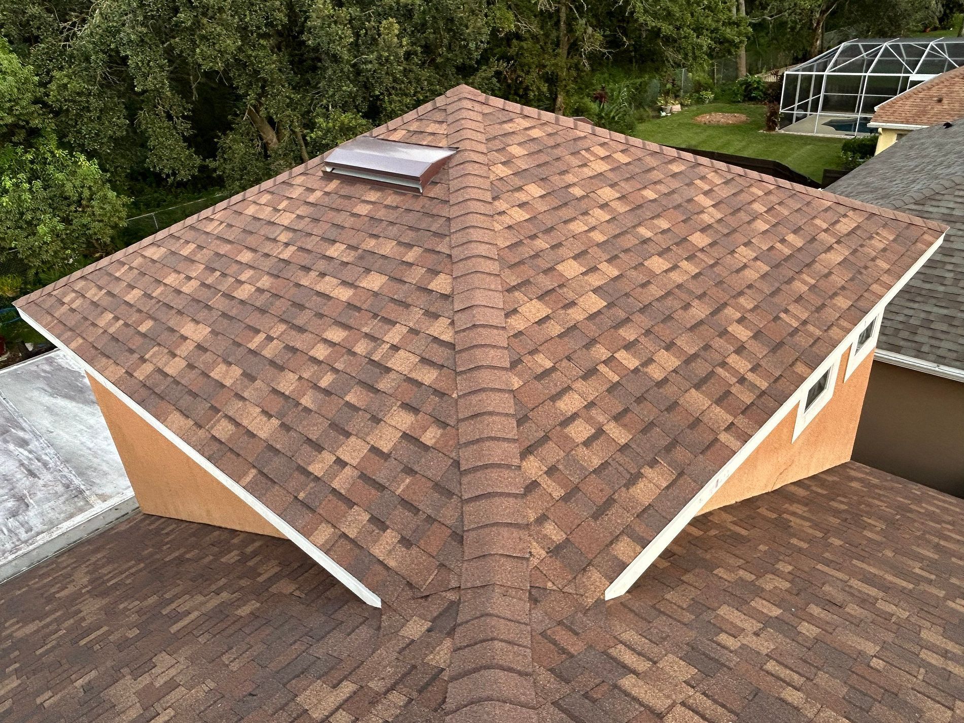 Brown shingled roof with a skylight, viewed from above, surrounded by trees and houses.
