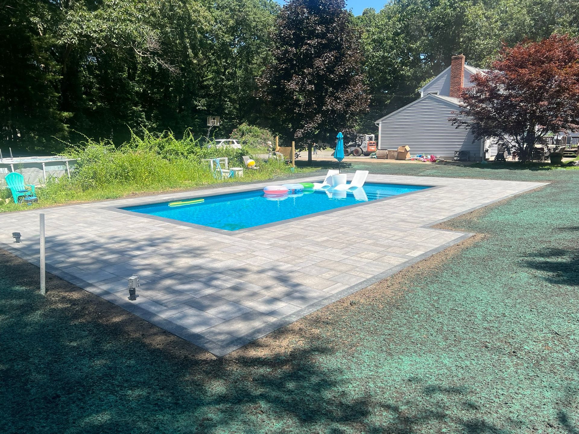 Rectangular pool surrounded by stone patio, green lawn, trees, and house on a sunny day.