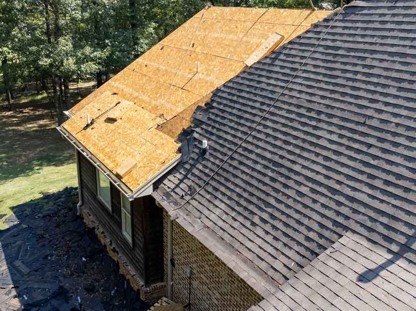 Roof under repair with exposed orange sheathing beside an older dark shingled roof