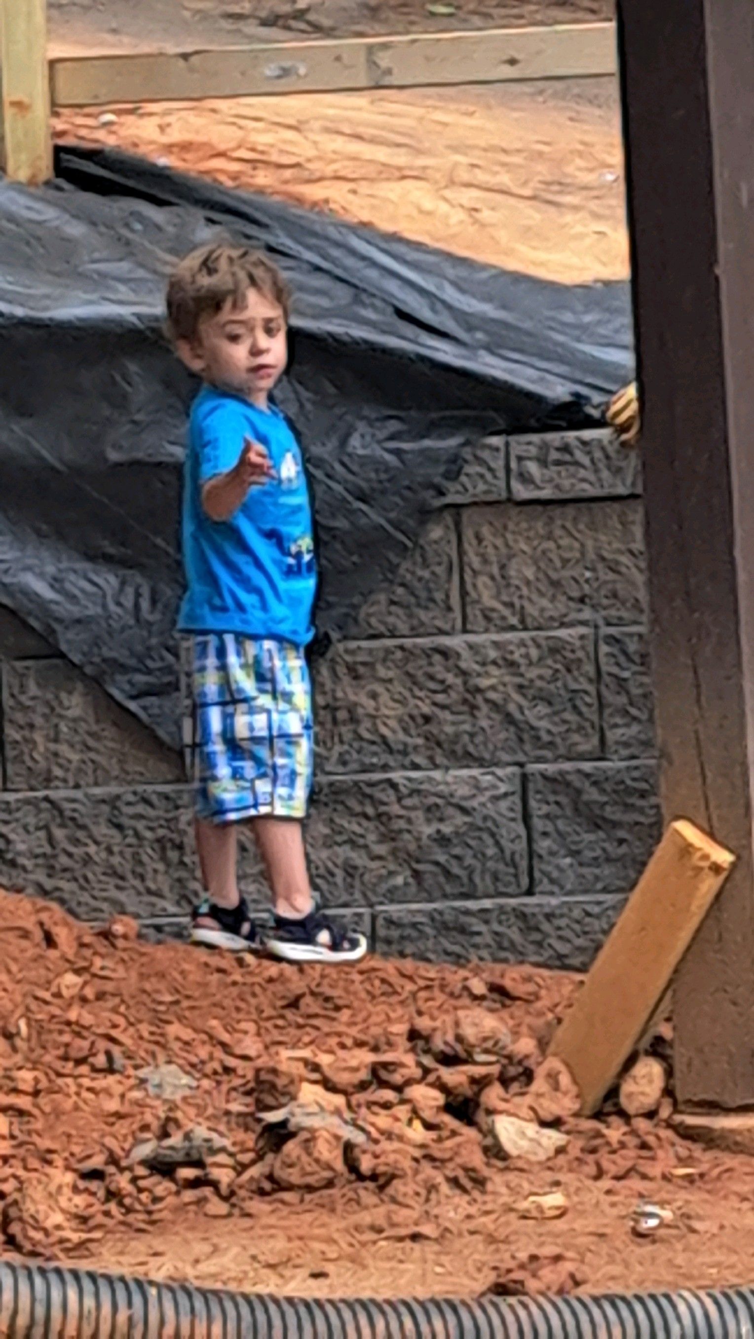 A young boy is standing in the dirt next to a brick wall.
