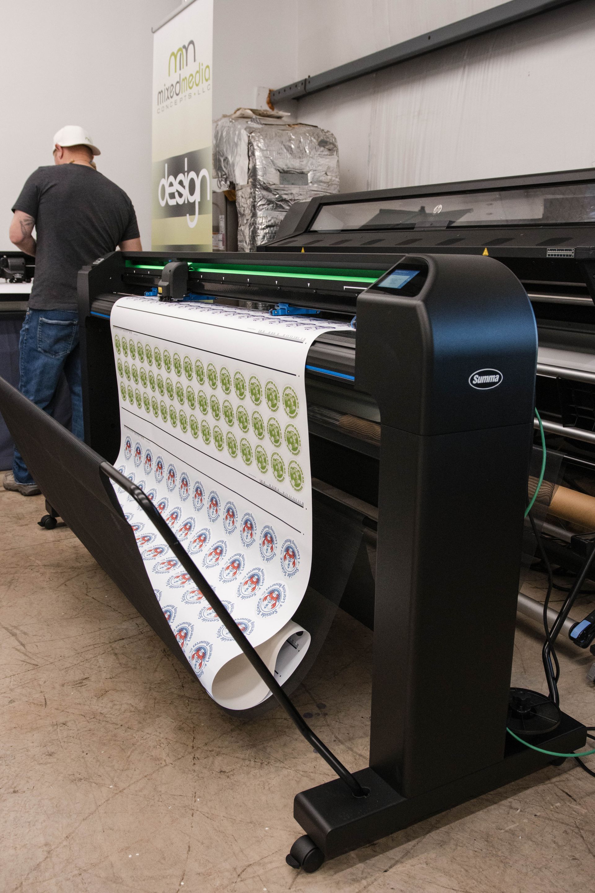 Large-format printer producing a long banner in a workshop, with a worker standing nearby.