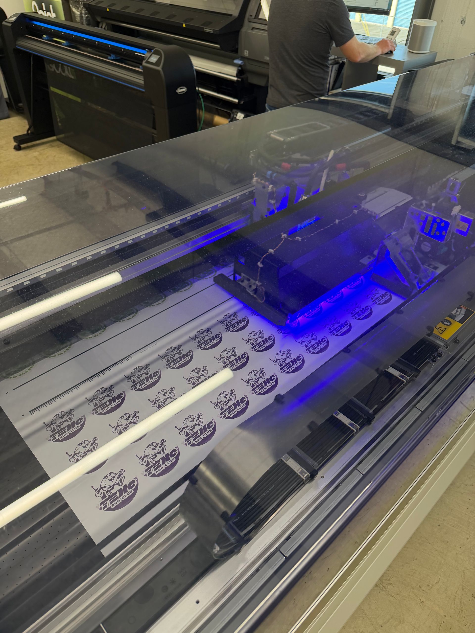 Large-format printer printing white sheets with black graphics under a blue UV light.