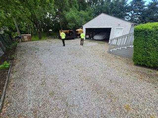 Two people are standing in a gravel driveway in front of a garage.