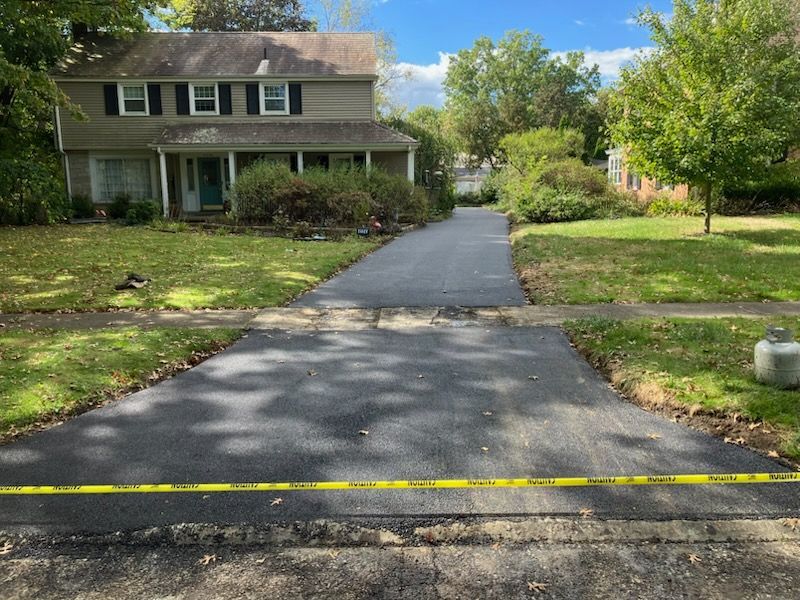 Newly paved asphalt driveway leading to a two-story house with green lawn. Yellow caution tape is across the road.