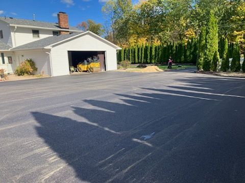 Asphalt driveway with garage, house, and tall green trees casting shadows on a sunny day.