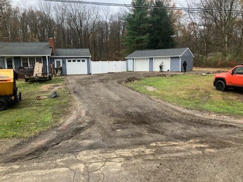 Driveway leading to blue-gray houses and garages; a red truck and equipment are on the side, surrounded by trees.