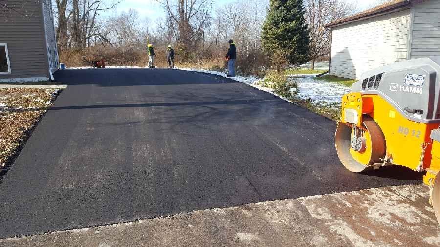 A roller is rolling asphalt in a driveway next to a house.