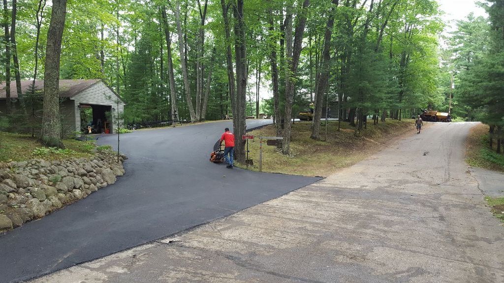 A man is riding a lawn mower down a road surrounded by trees.