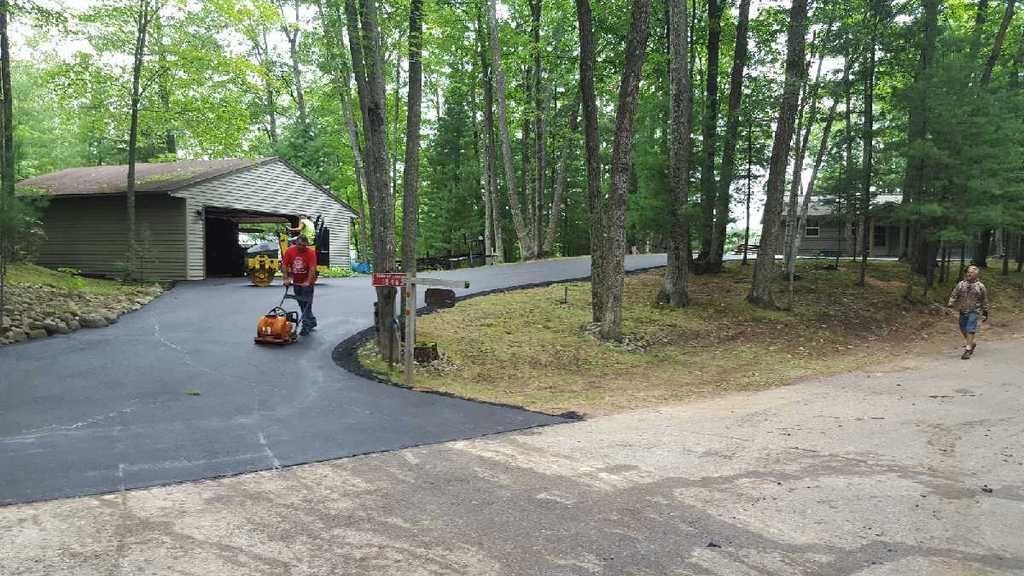 A man is pushing a lawn mower down a driveway surrounded by trees.