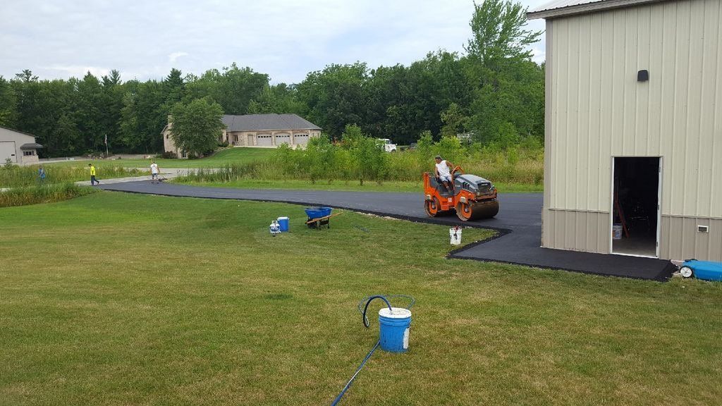 A roller is rolling asphalt on a driveway in front of a building.