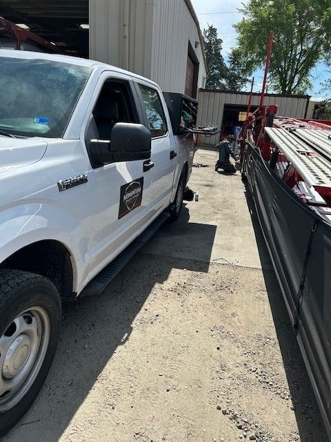 White utility truck parked in a narrow alley beside a building and dumpster, with ladders on a trailer ahead