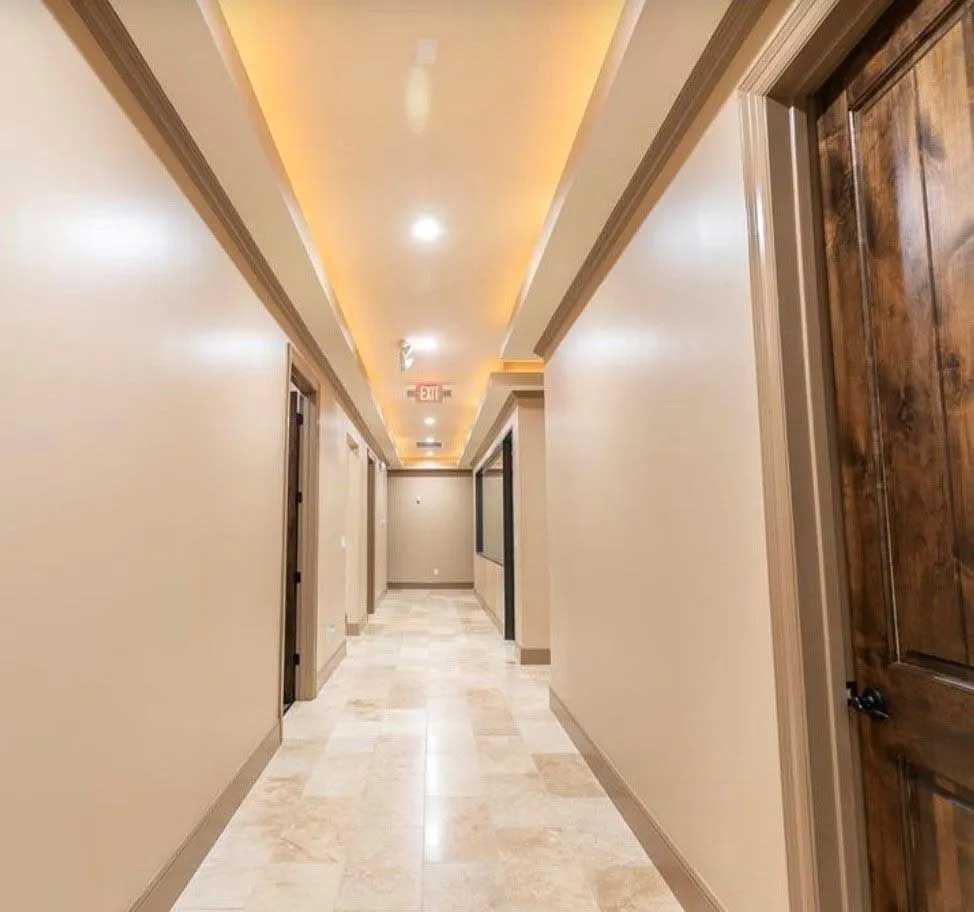 Long hallway with beige walls, tan tile floor, and recessed lighting in the ceiling. A wooden door is on the right.