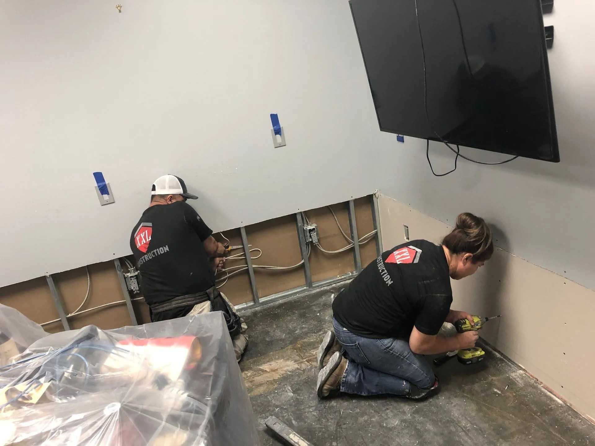 Two people installing drywall in a room with a mounted TV.