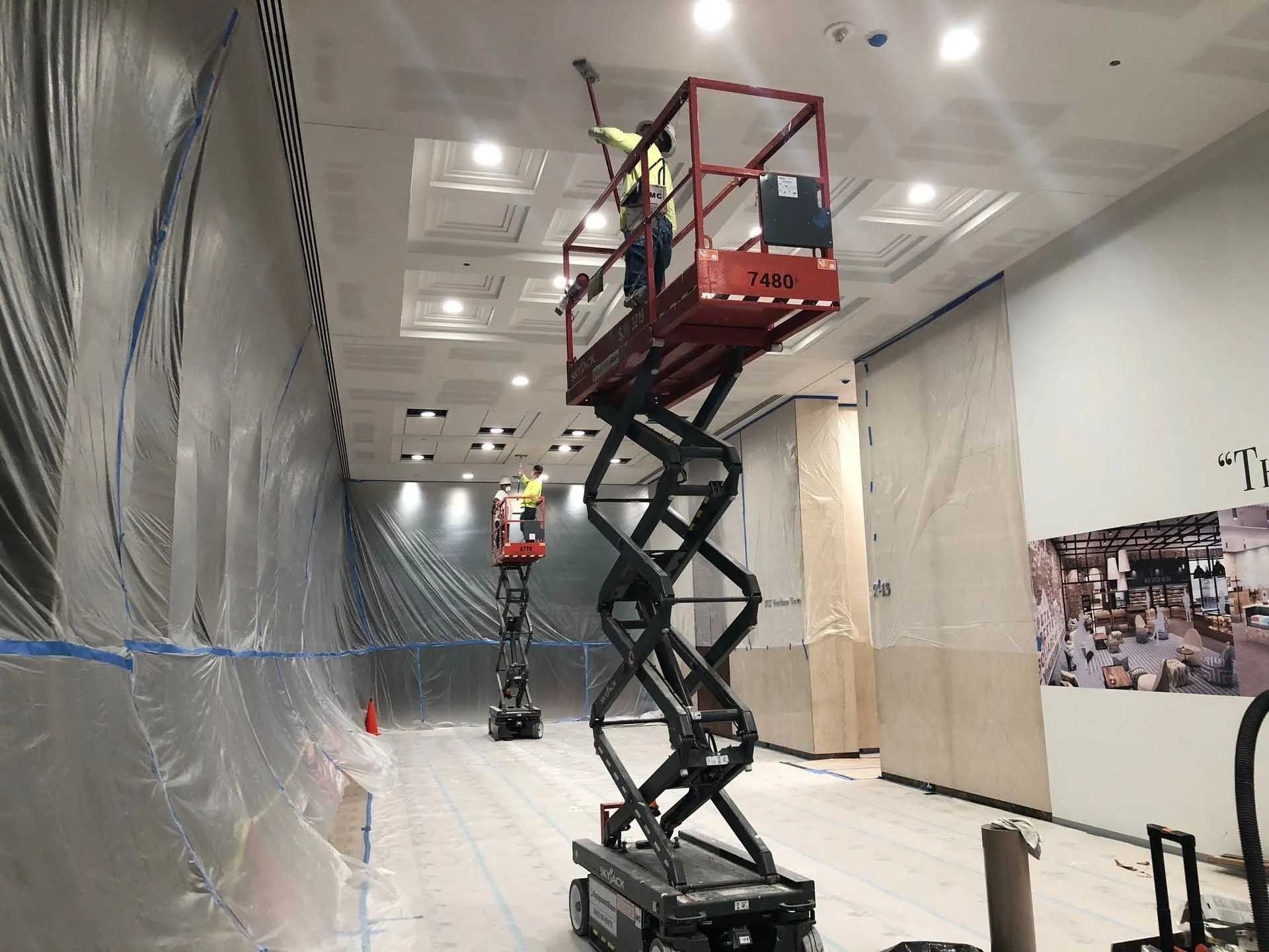 Workers on scissor lifts paint a ceiling in a room with plastic sheeting and partially covered walls.