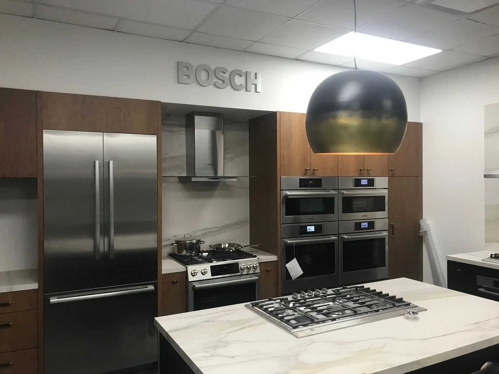 Kitchen appliance showroom with Bosch logo; stainless steel refrigerator, oven, stove, and island with cooktop.