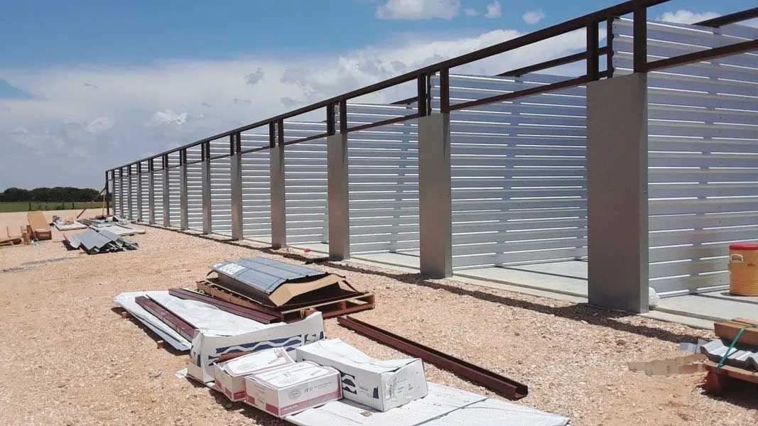 Construction of a long, corrugated metal and concrete wall under a blue sky, with building materials in foreground.