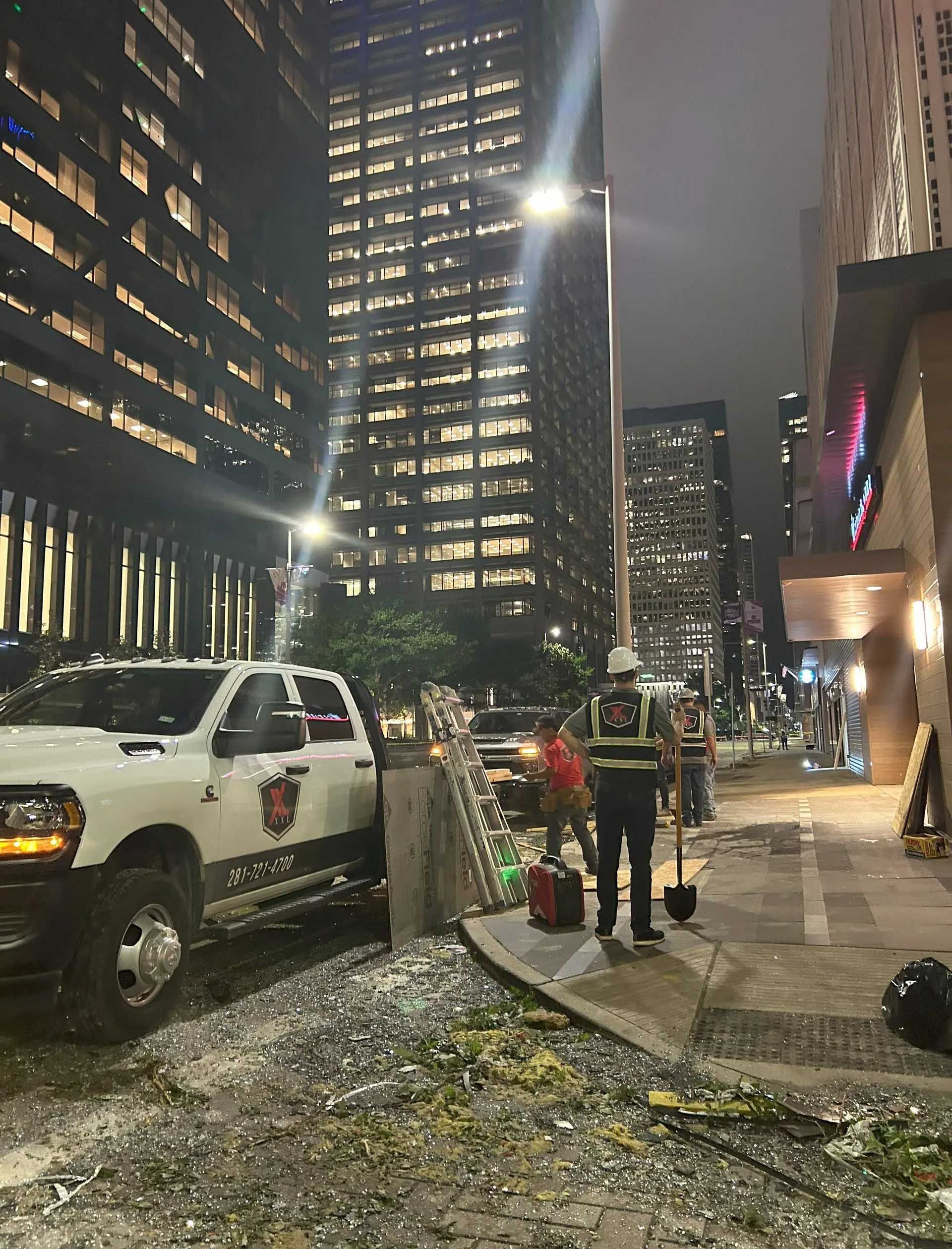 Construction workers near a white truck on a city street at night. Tall buildings and streetlights are in the background.