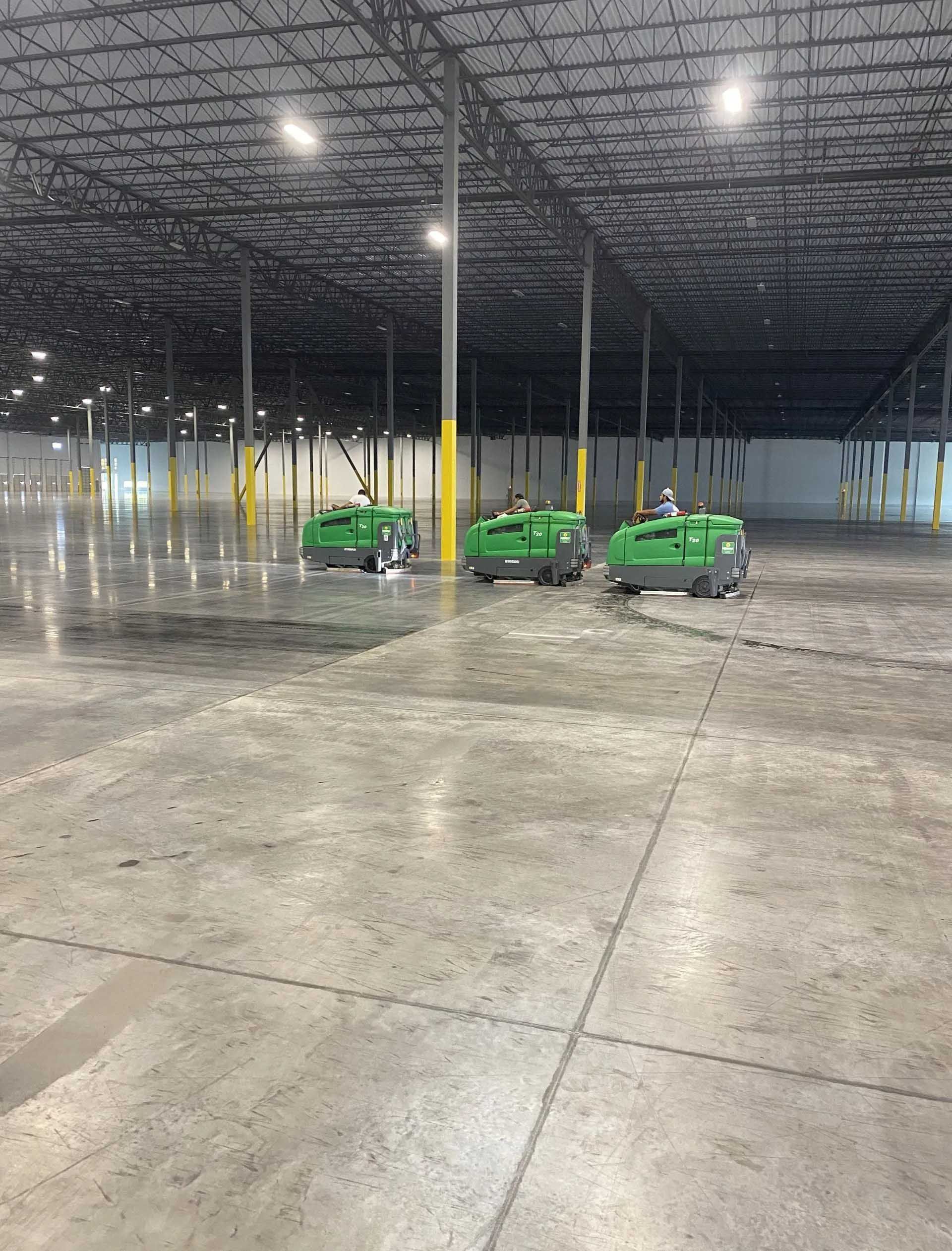 Three green floor cleaning machines operating in a large, empty warehouse.