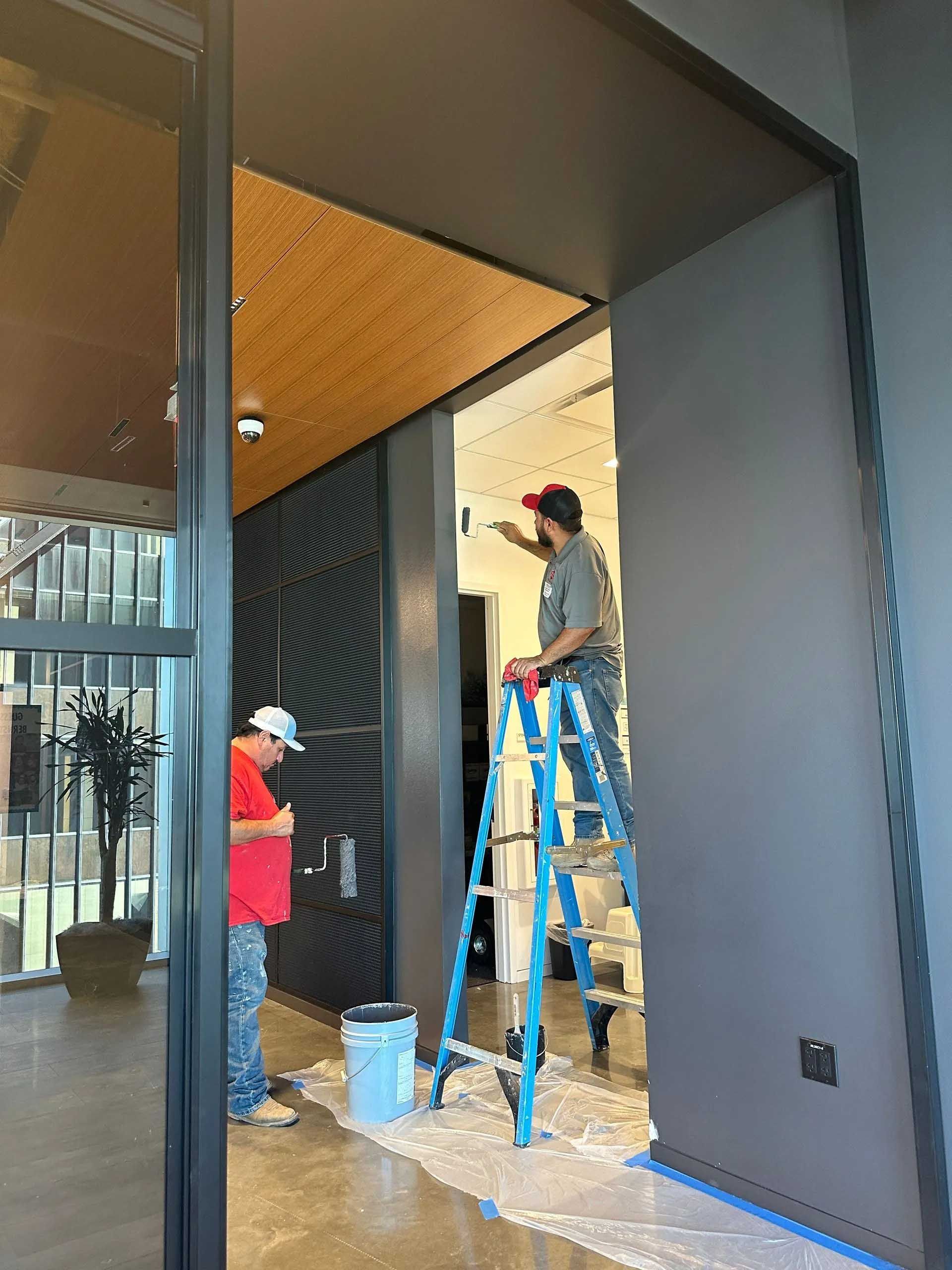 Two workers painting a doorway interior. One on ladder, other standing nearby. Gray walls, wood ceiling.
