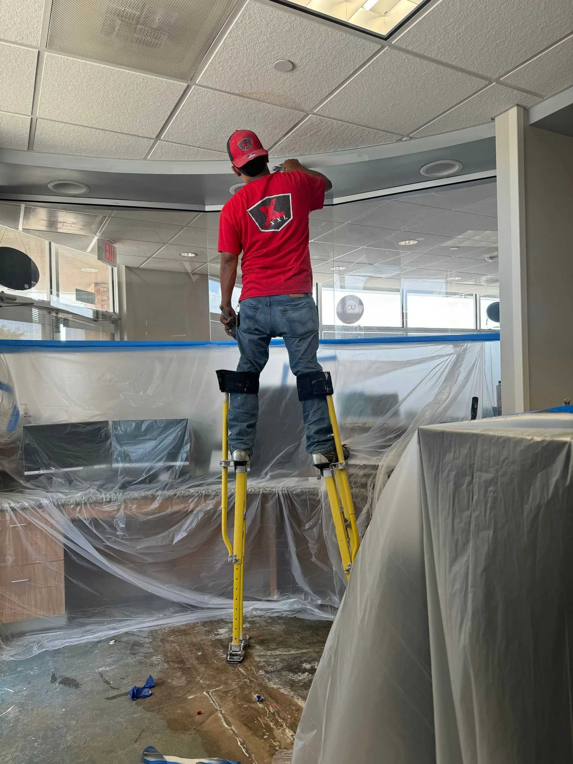 Person in red shirt and jeans on stilts, painting a ceiling indoors.