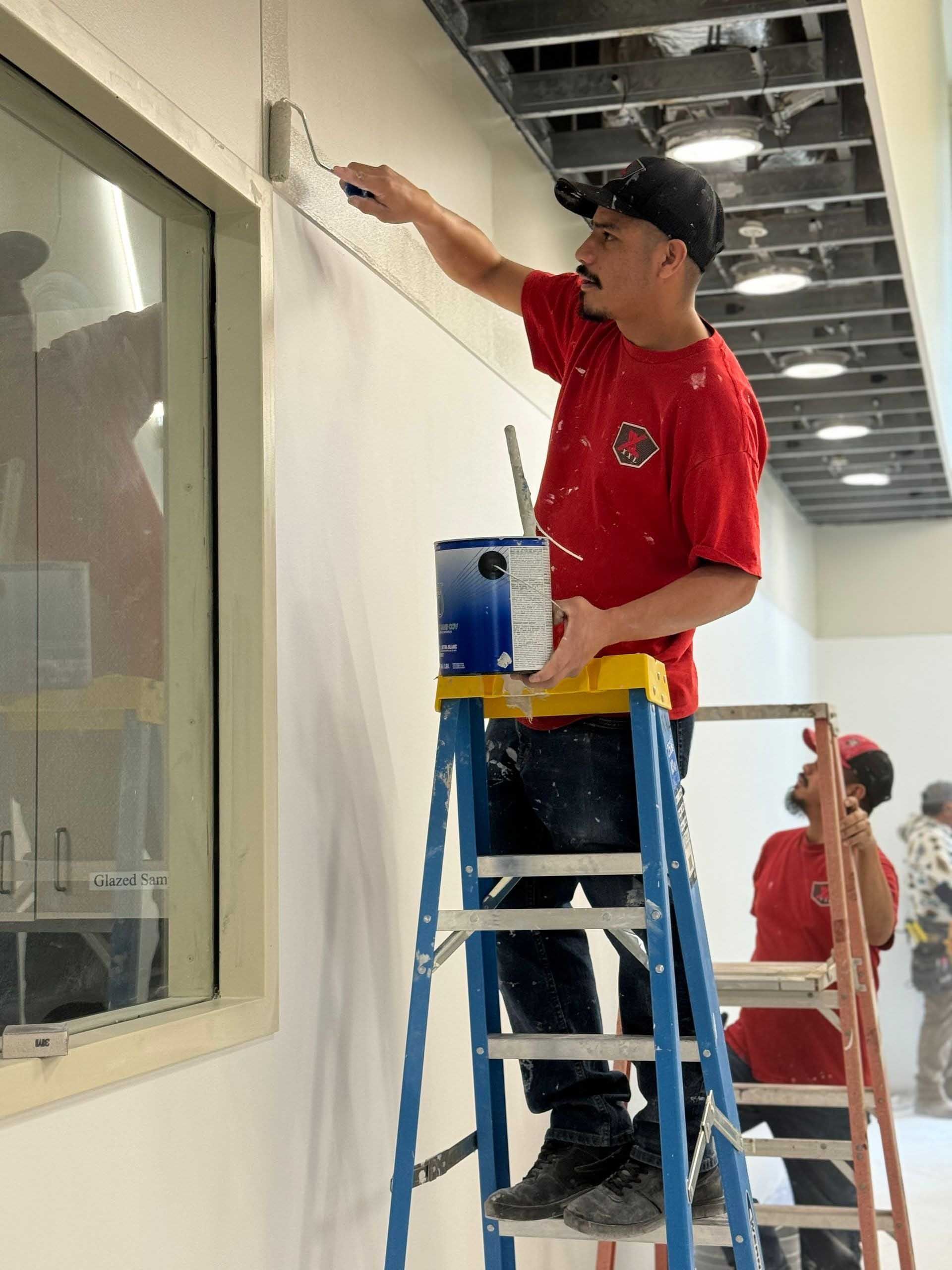 Two people painting an interior wall. One on a ladder, rolling paint on the wall. The other, also on a ladder, out of focus.