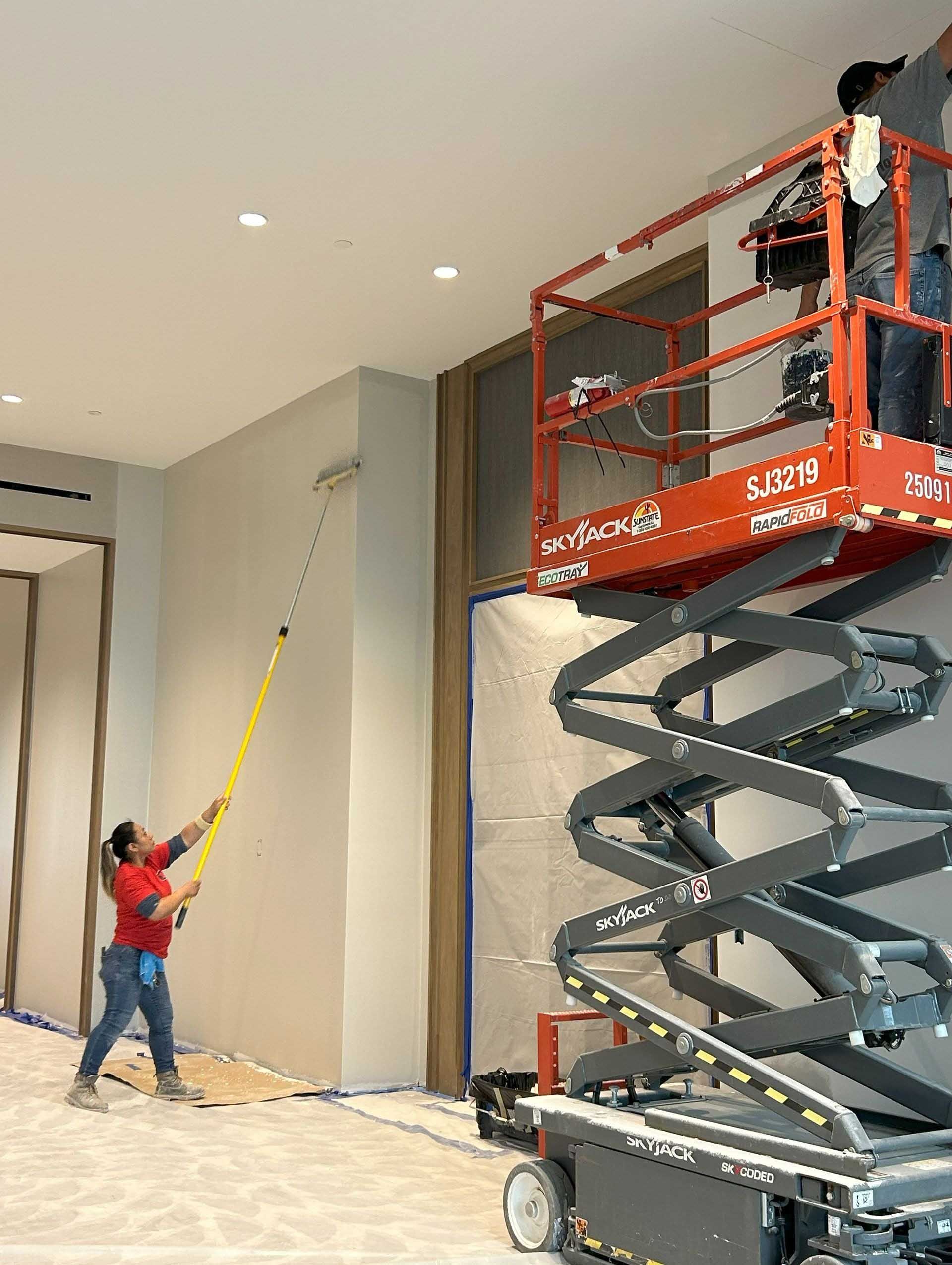 Two people painting a ceiling, one on a lift, the other with a pole. Indoors, construction site.
