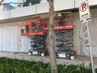 Workers on scissor lifts beside a building, near a parking sign.