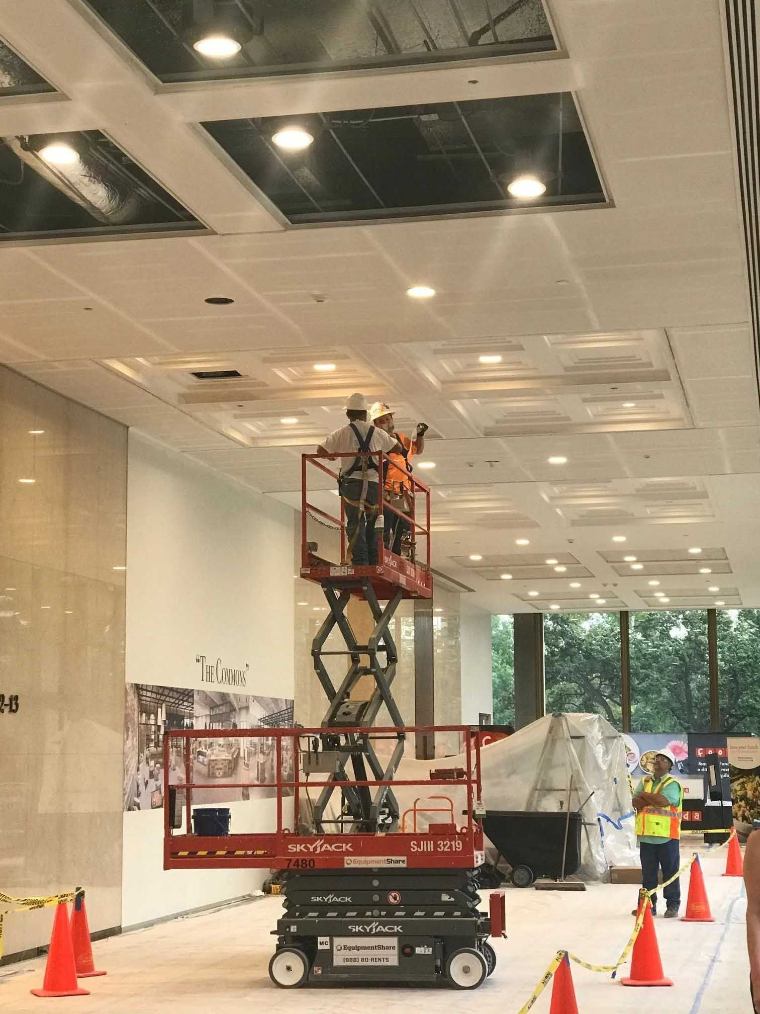 Construction workers on a lift installing ceiling panels, indoors. Safety cones and temporary barriers present.