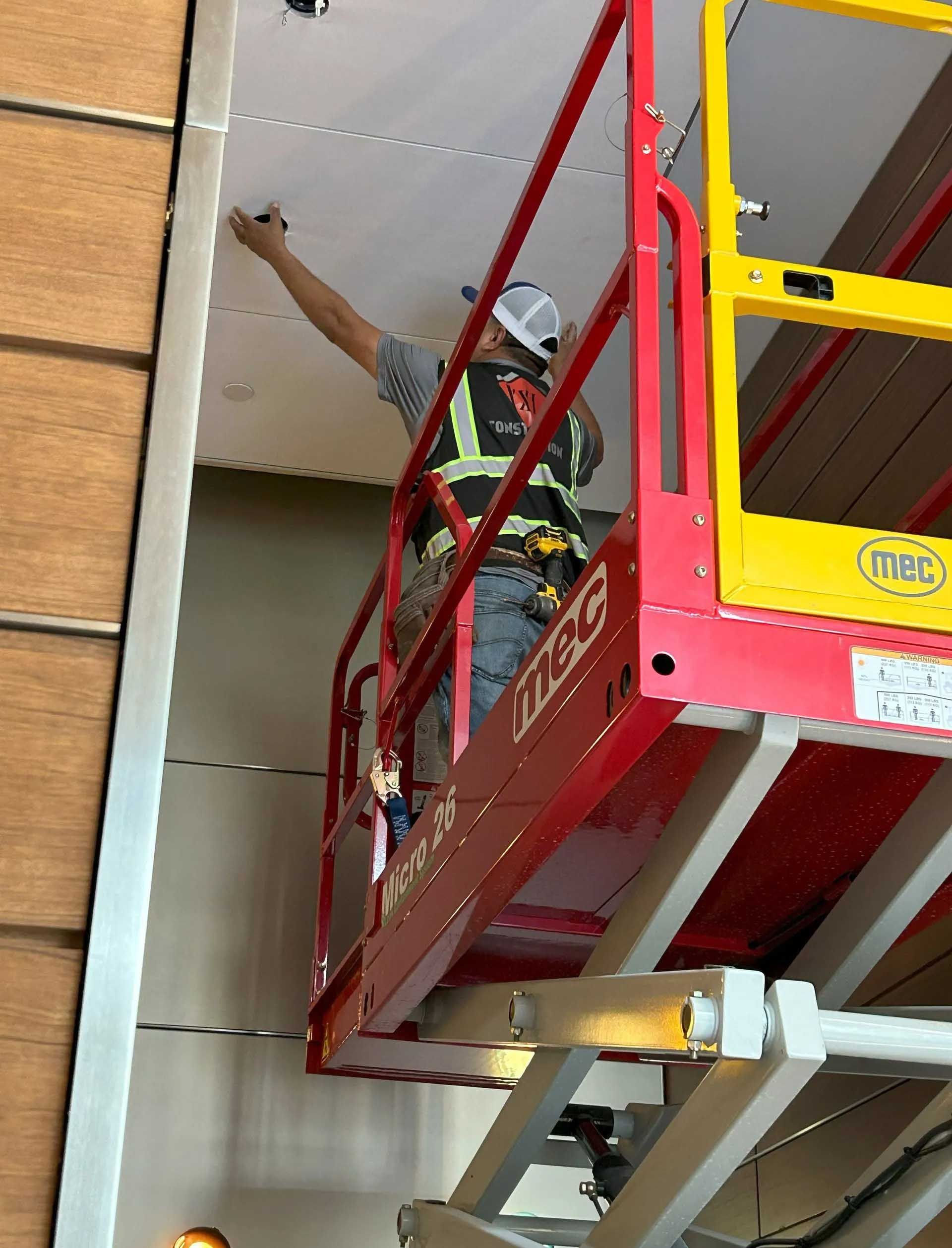 Construction worker on a scissor lift installs something on a white ceiling. Wooden wall to the left.