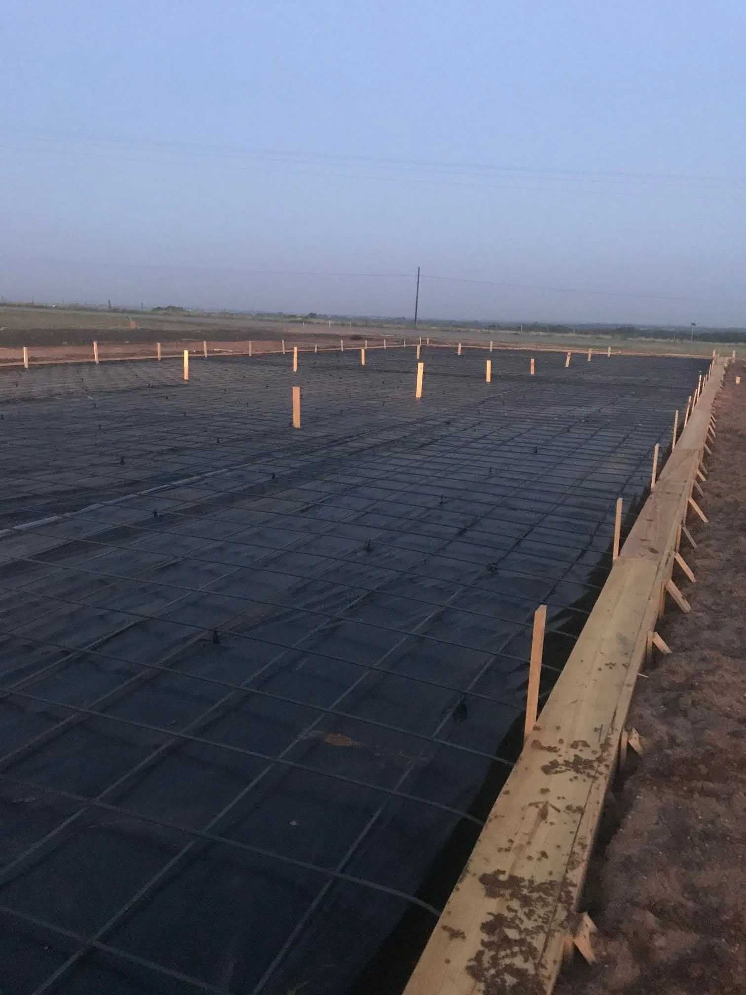 Construction site: a rectangular form with rebar grid and black tarp, wooden border, stakes.