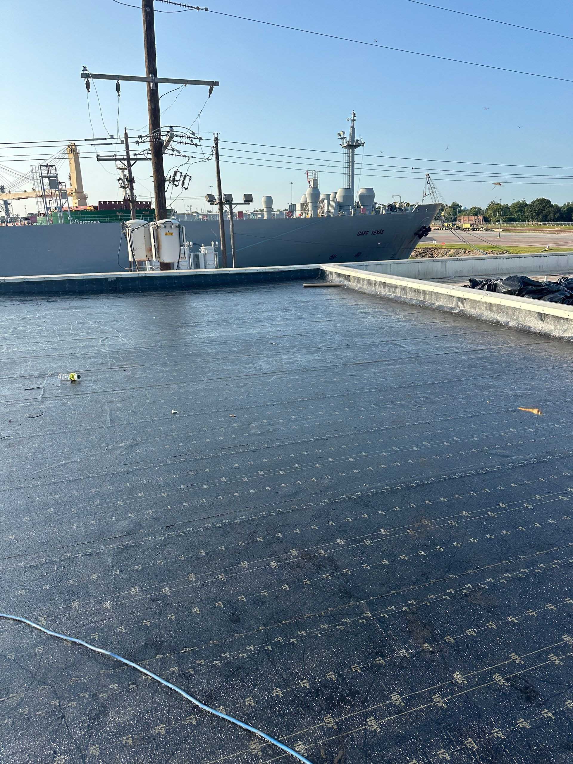 A dark, flat roof with a large ship docked in the background on a sunny day.