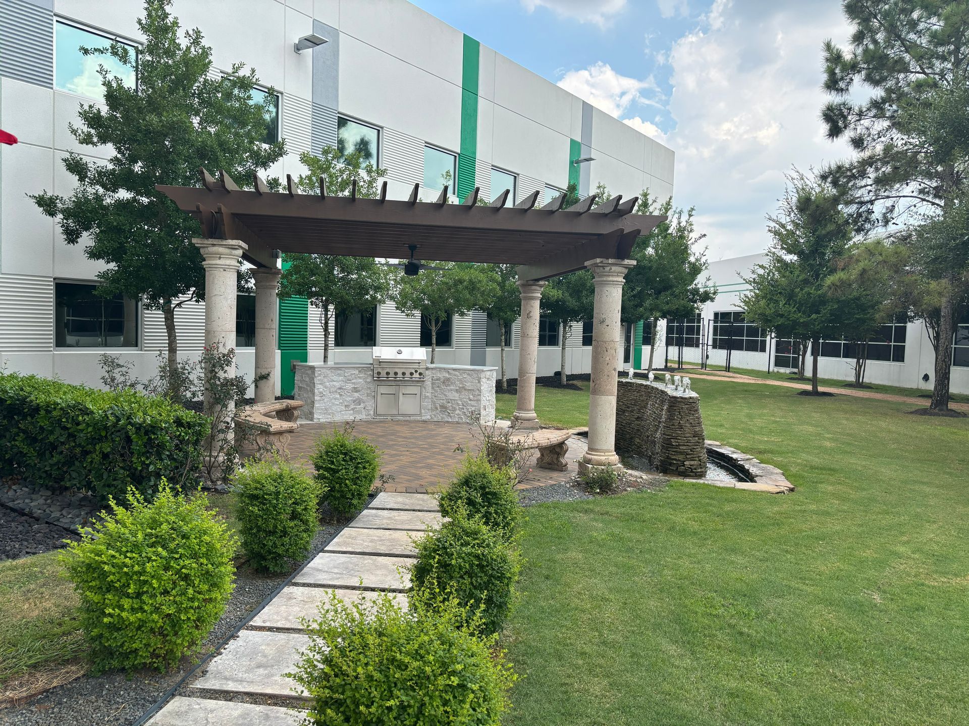 Stone pergola with columns in a grassy outdoor area in front of a white building with green accents.
