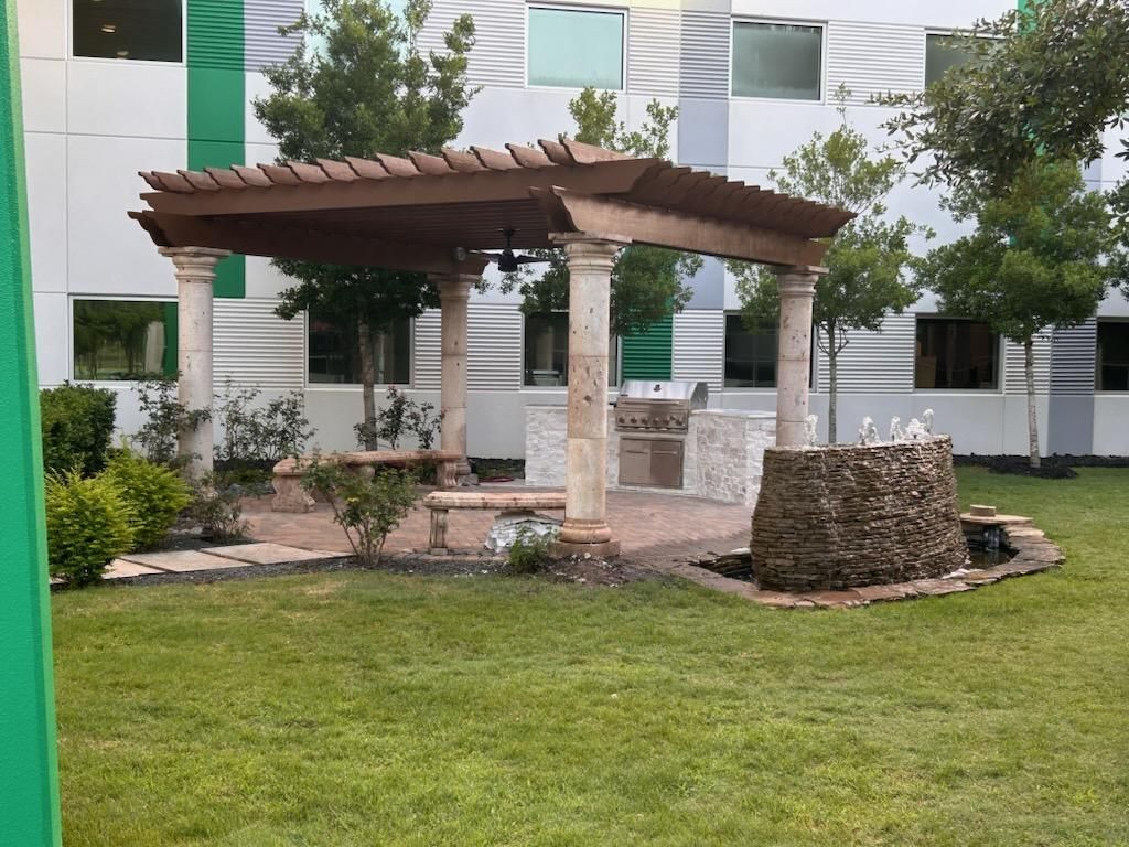 Pergola with columns, grill, fountain, and seating on a brick patio in a grassy area next to a building.
