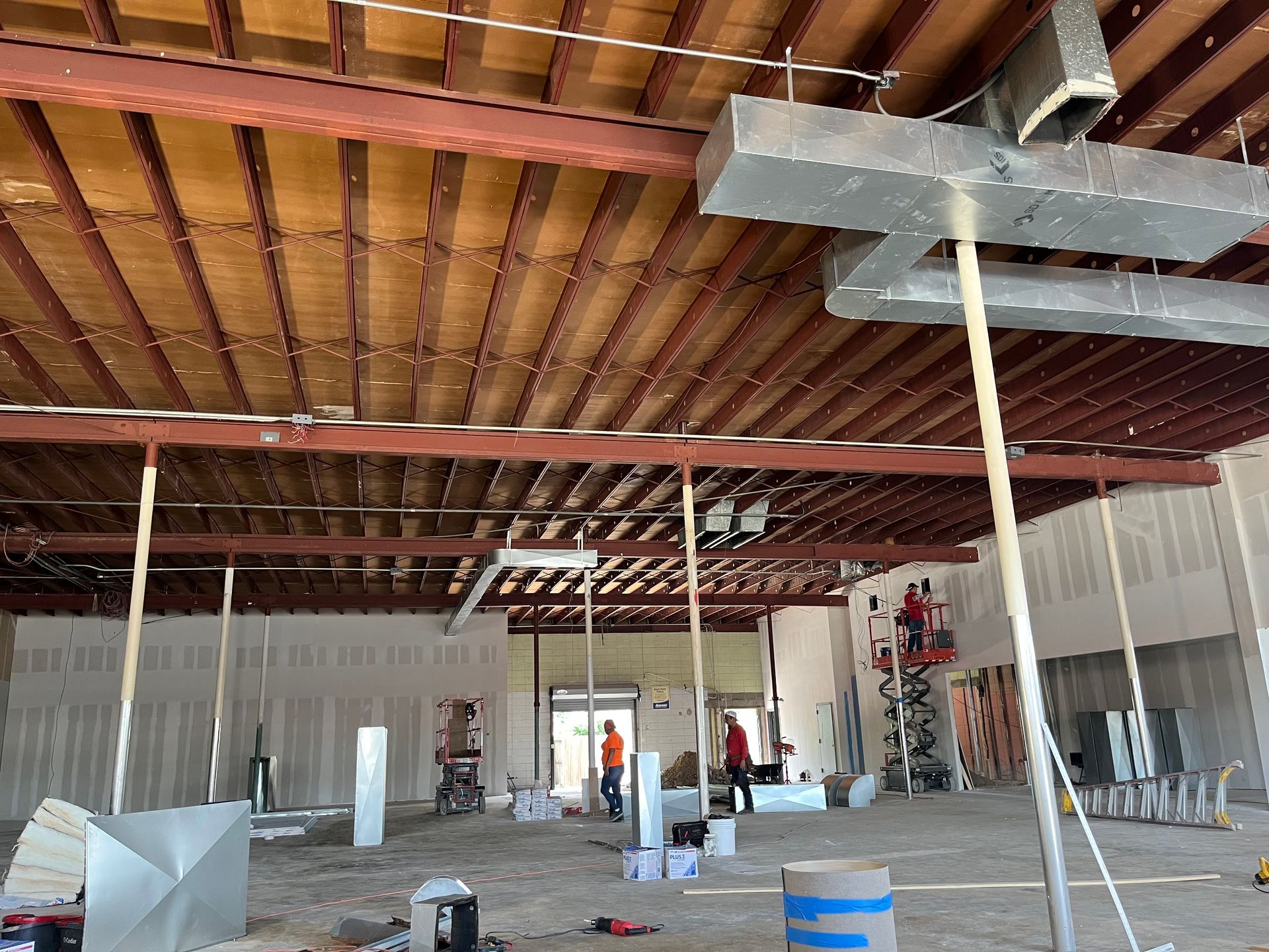 Interior of a construction site with exposed ceiling beams, HVAC ductwork, and workers.