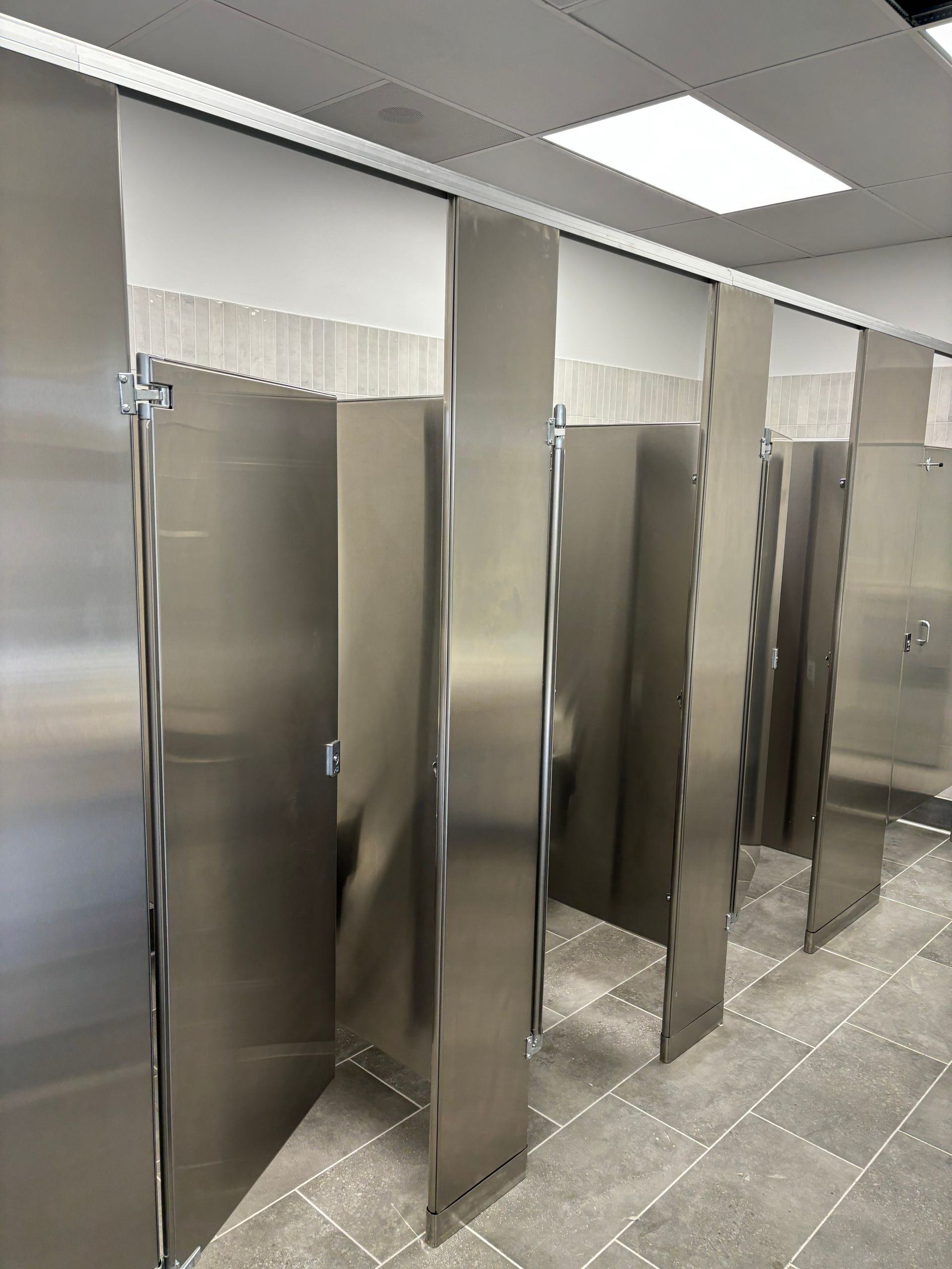 Stainless steel restroom stalls in a public restroom with grey tile flooring.