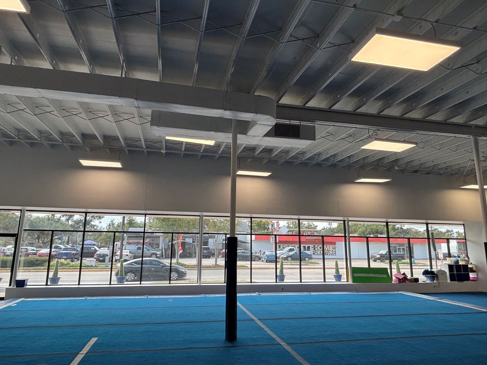 Blue mat in a gym with windows. Metal ceiling with rectangular lights. Black pole visible.