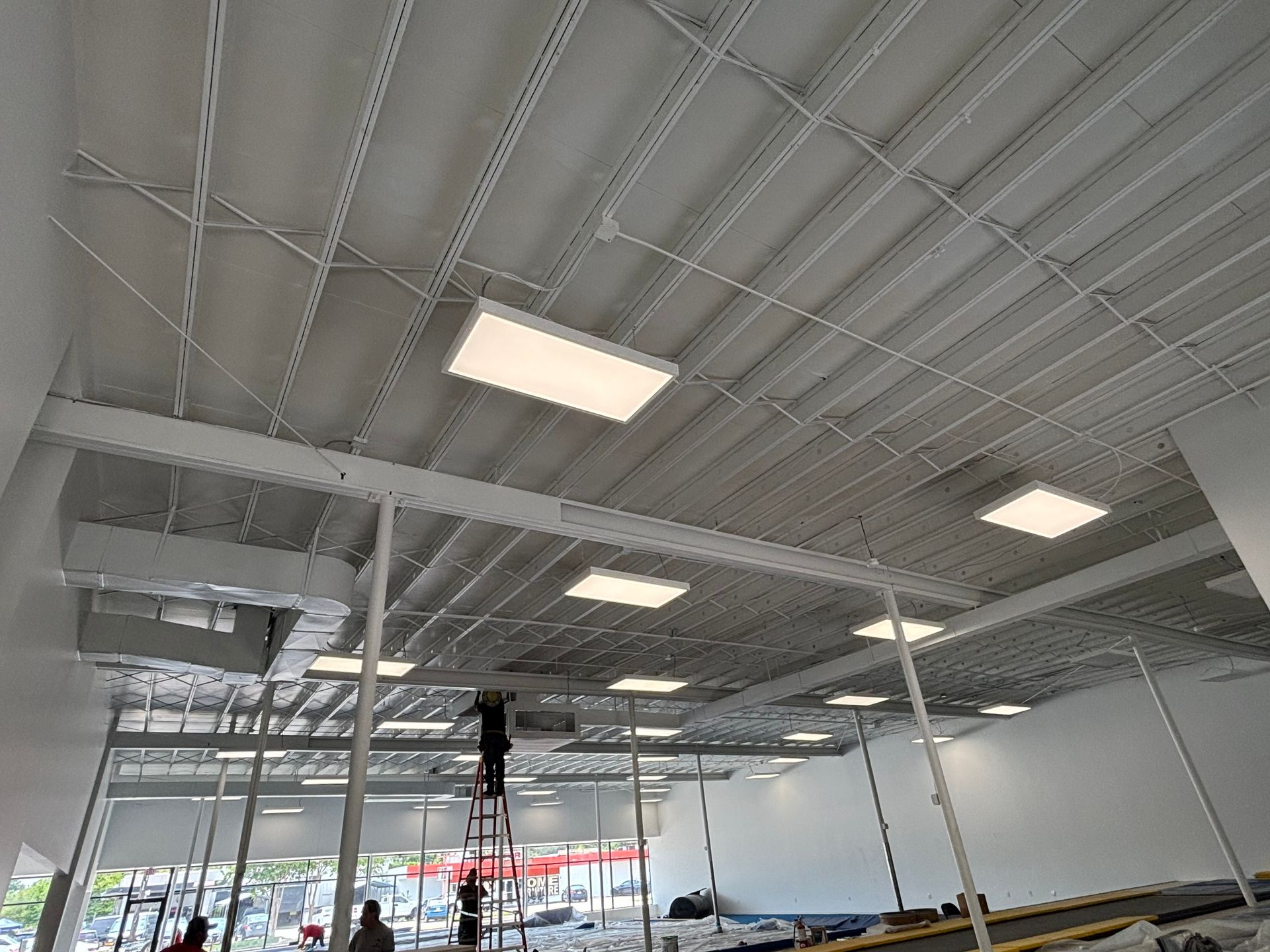 White interior ceiling with rectangular lights, beams, and vents; people on a ladder.