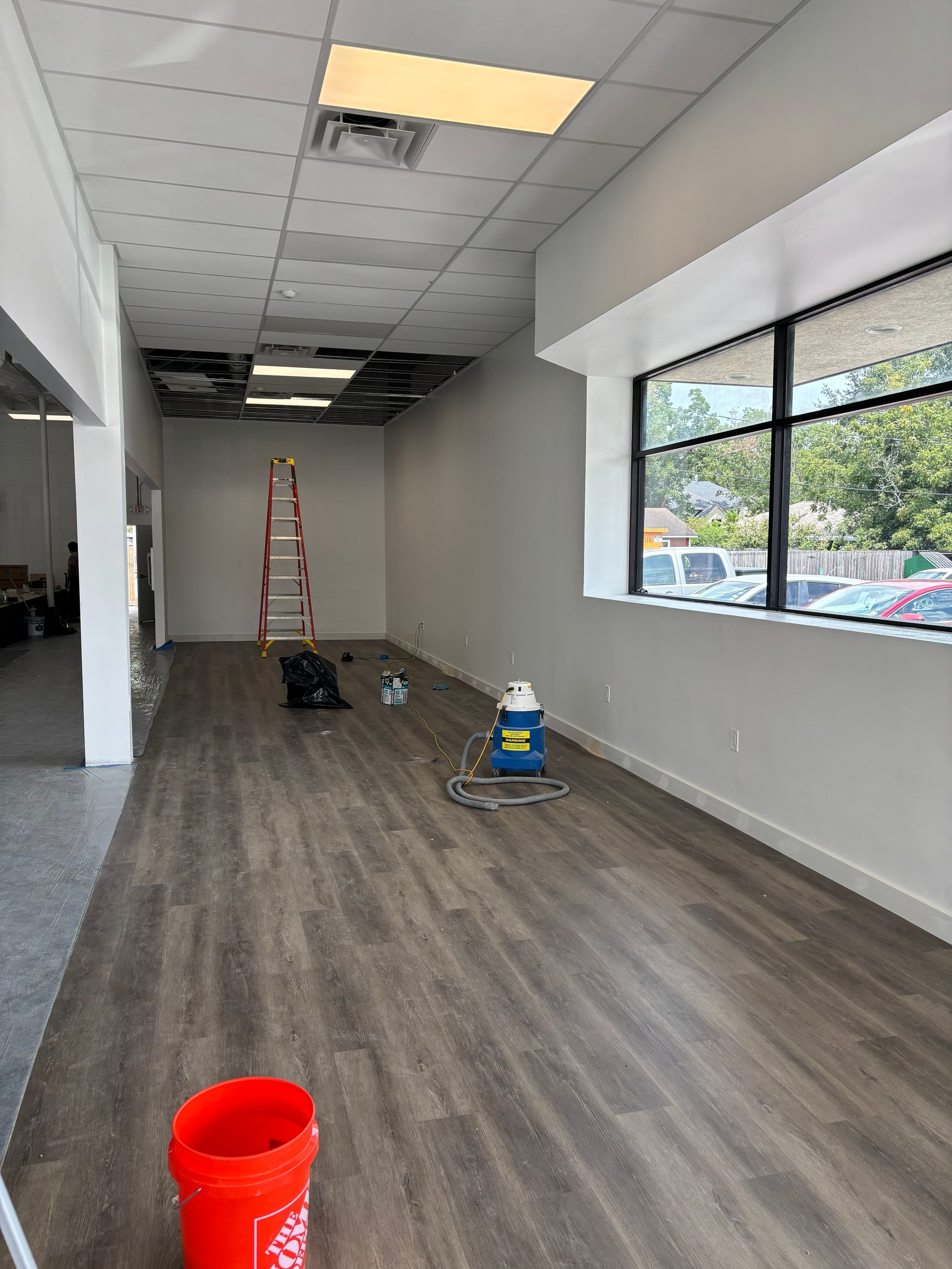Interior of a retail space under construction, featuring exposed ceiling tiles, wood-look flooring, and a window.