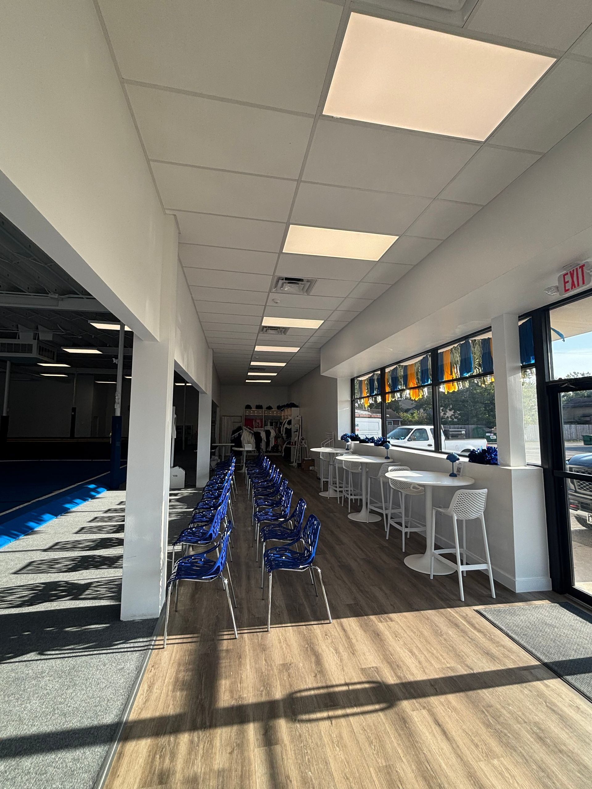 Interior of a brightly lit space with blue chairs, white tables, and large windows.