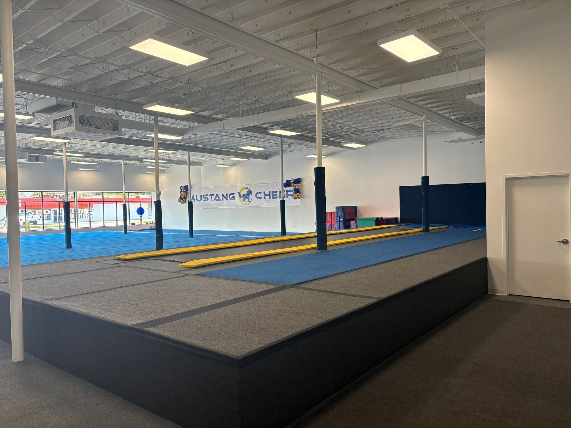 Gymnasium interior with blue and gray mats, black platform, white poles, and ceiling lights.