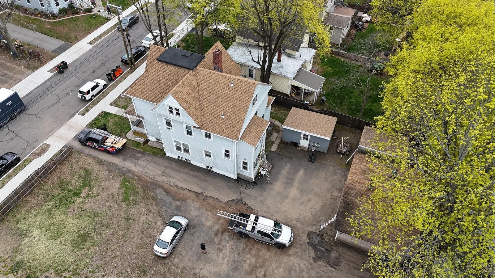 Aerial view of a white house with a tan roof, parked cars, and surrounding trees and dirt lot.