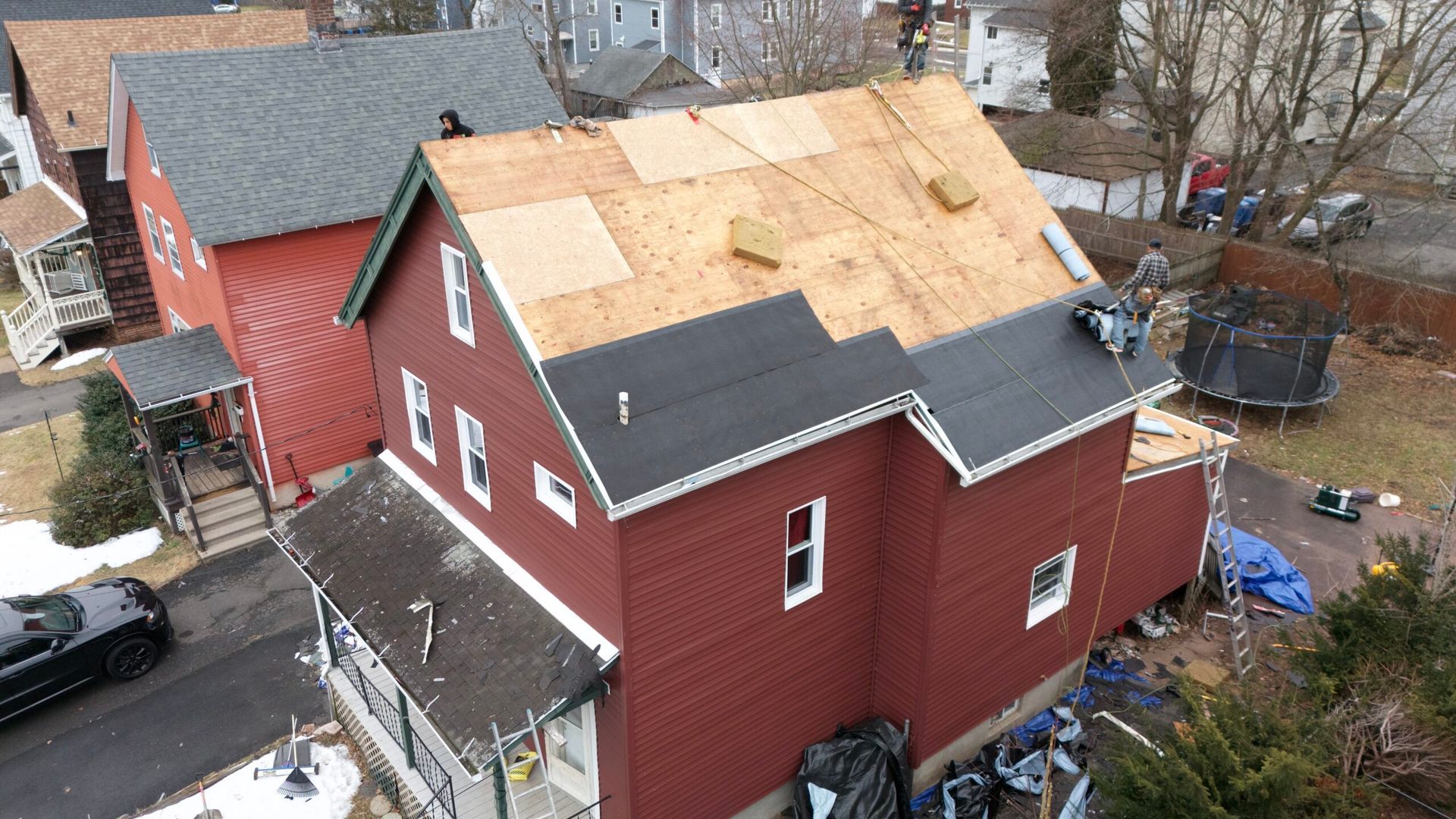 Aerial view of a red house with a partially replaced plywood roof in a residential neighborhood.