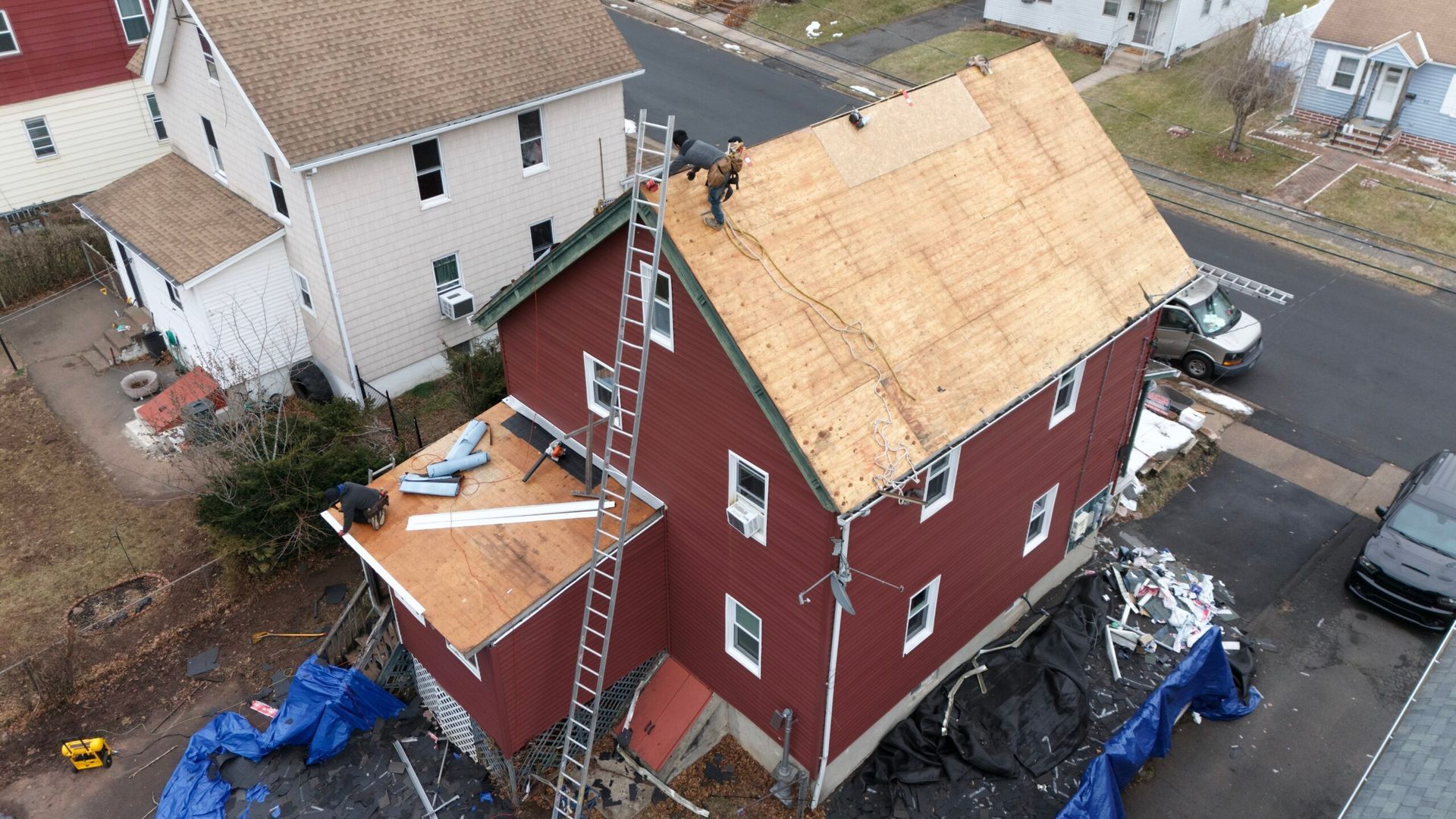 Aerial view of a red house under roof repair, with plywood sheathing, ladders, and blue tarps around it