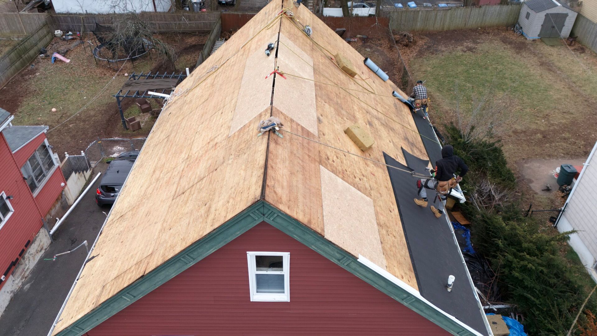 Aerial view of a house roof under repair, with exposed plywood and workers on the right side.