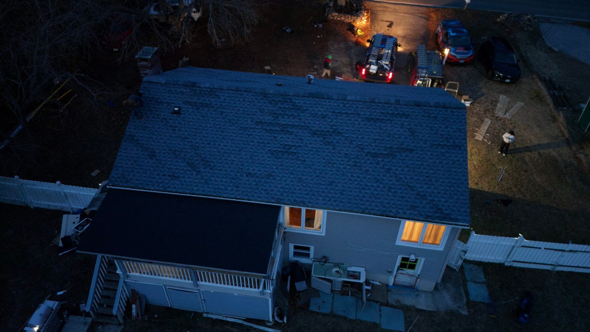 Aerial view of a house at night with a blue roof and warm lights glowing inside, surrounded by darkness.