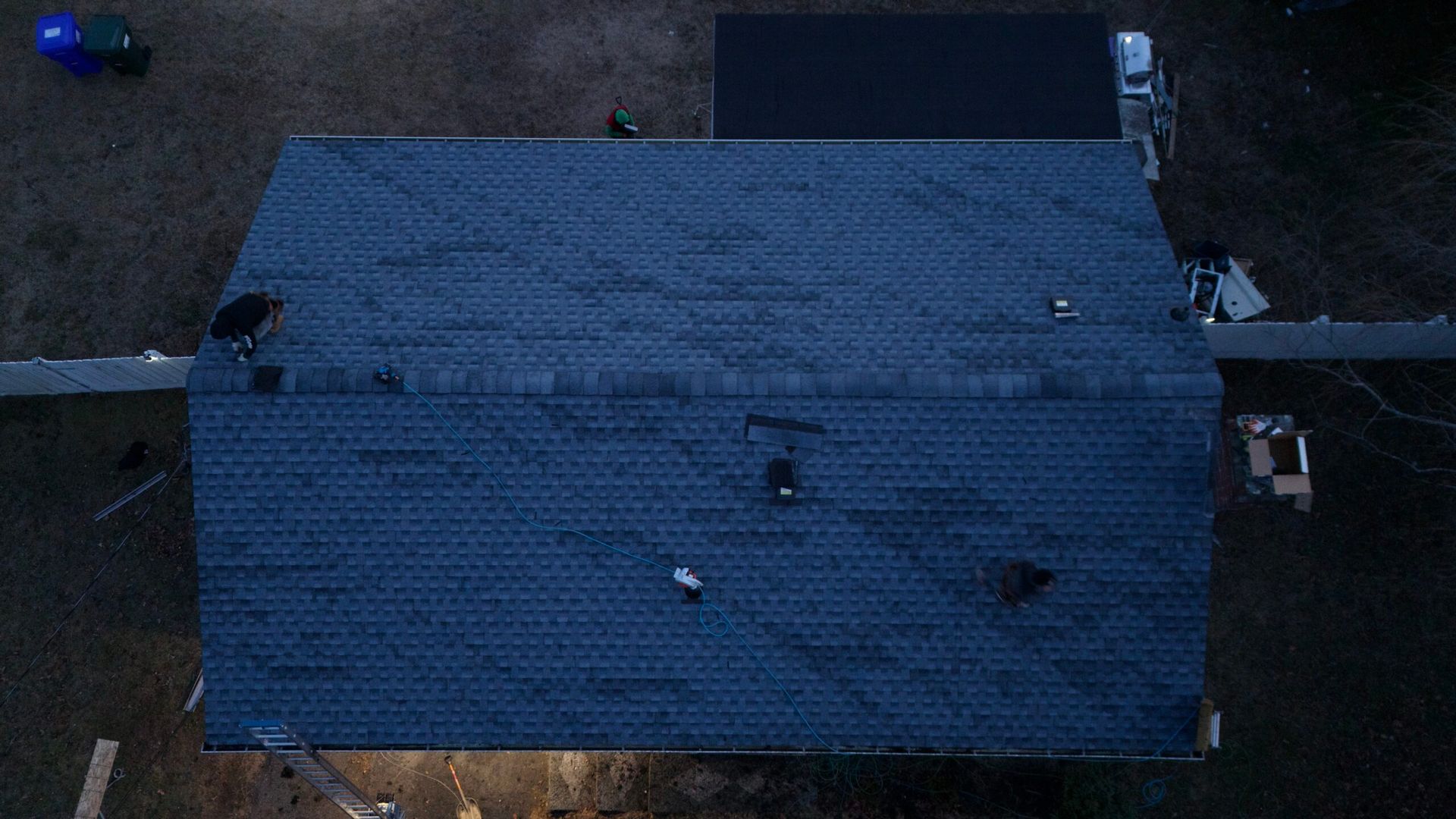 Aerial view of a blue-roofed house with a few people or objects around it on a dark lot.
