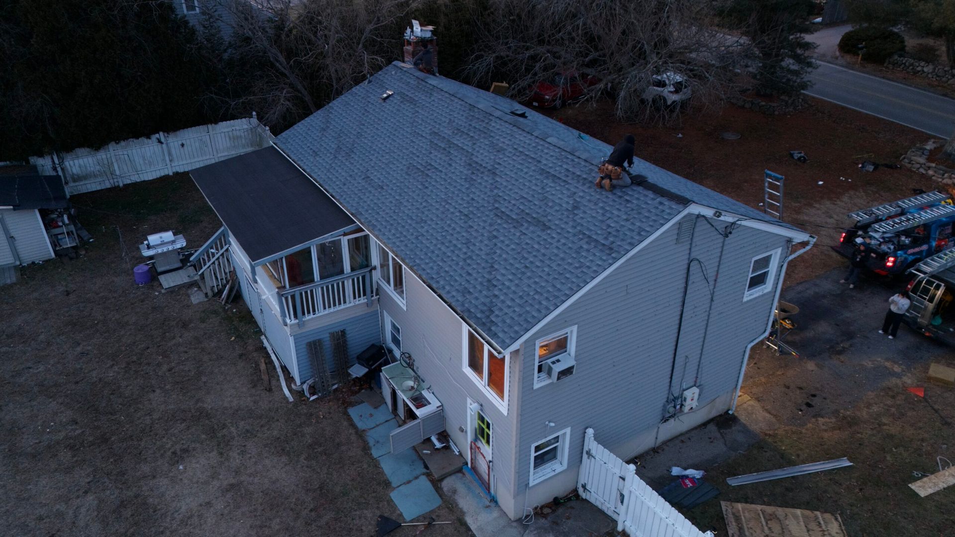 Aerial view of a gray house with a dark roof, porch, and cars parked in the driveway at dusk