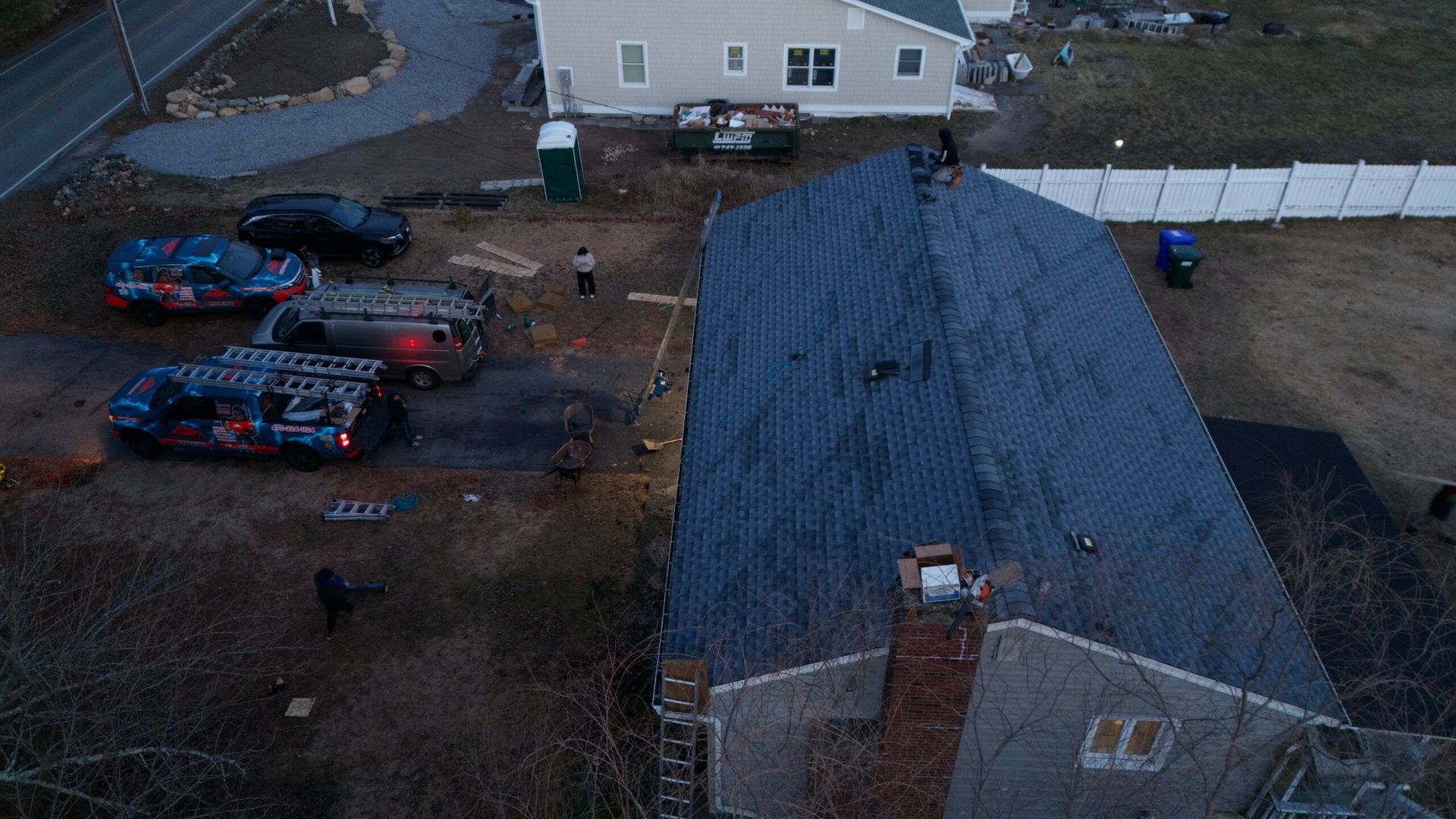 Aerial view of a house with a dark roof, parked cars, and a fenced yard at dusk.