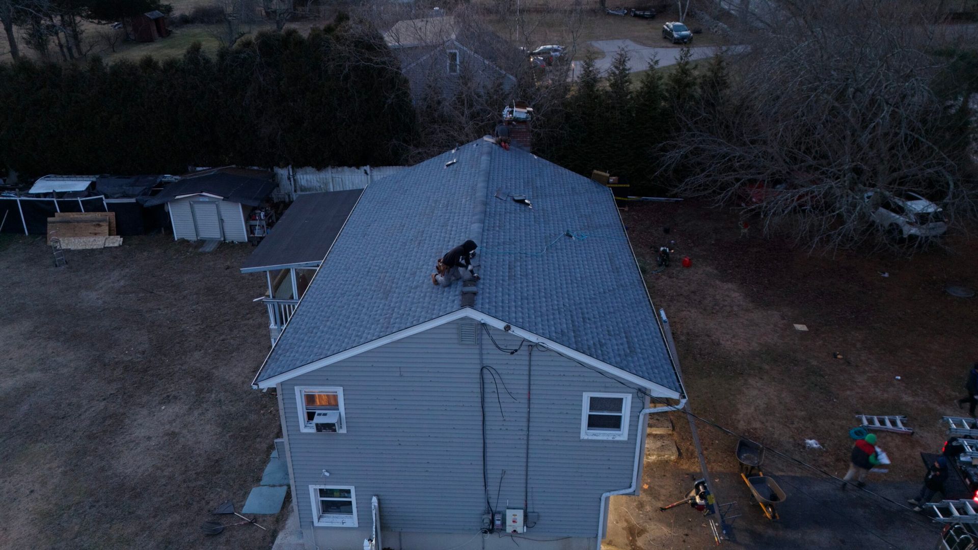 Aerial view of a gray house roof with a person working on it, surrounded by trees and yard equipment.